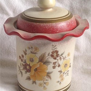 May include: A white ceramic cookie jar with a red and gold rim and a floral design. The lid has a scalloped edge and a knob on top.