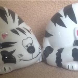 May include: Two ceramic sleeping cat figurines. The cats are white with black stripes and have pink noses and black eyes. They are curled up in a sleeping position.