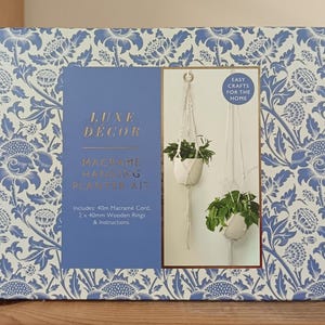 May include: A macrame hanging planter kit in a box with a blue and white floral design. The kit includes macrame cord, wooden rings, and instructions. The box shows two hanging planters with plants. The text reads 'Easy Crafts for the Home'.