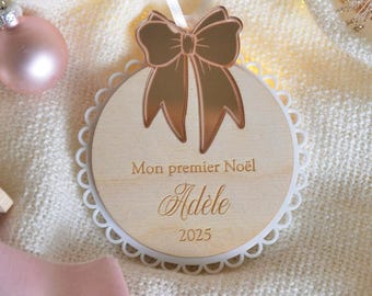 Könnte beinhalten: Ein hölzerner Weihnachtsschmuck mit einer goldenen Schleife und dem Text "Mon premier Noël Adèle 2025". Der Schmuck hat einen weißen, gewellten Rand und wird von einem weißen Band gehalten. Zwei rosa und weiße Weihnachtskugeln sind ebenfalls zu sehen.