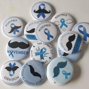 Op de afbeelding: Een verzameling witte, ronde buttons met blauwe accenten. Elke button toont het woord "MOVEMBER" en diverse ontwerpen, waaronder snorren, linten en zinnen als "ON GARDE LA MOUSTACHE". De buttons zijn waarschijnlijk gerelateerd aan bewustwording van mannengezondheid.