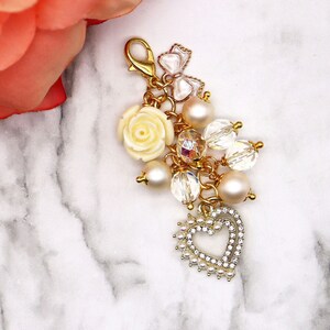 May include: A gold charm with a white rose, pearls, and crystals. The charm is attached to a gold chain and has a heart-shaped pendant.