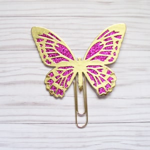 May include: A gold-colored butterfly paper clip with magenta glitter-filled wings. The butterfly design has intricate cut-out patterns. The paper clip is made of metal and is designed to hold papers together.