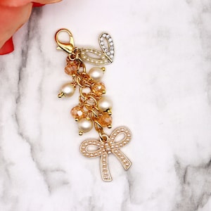 May include: A gold charm with a pearl and crystal embellishment, a gold chain, and a pearl bow. The charm is attached to a gold loop.