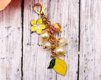 May include: A gold-colored charm with a yellow flower, faux pearls, and a yellow lemon pendant. The charm also features iridescent beads and a lobster clasp for attachment. The background is a light wood grain.
