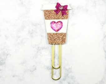 May include: A decorative paperclip in the shape of a coffee cup. The cup is covered in gold glitter with a white band and a pink heart. A pink glitter bow is at the top, and the clip is gold.