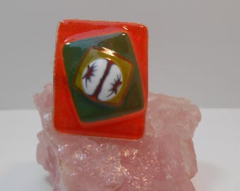 May include: A colorful ring with a rectangular orange base. The ring features a green and yellow square design with a white oval and red starburst details. The ring is displayed on a pink rock.