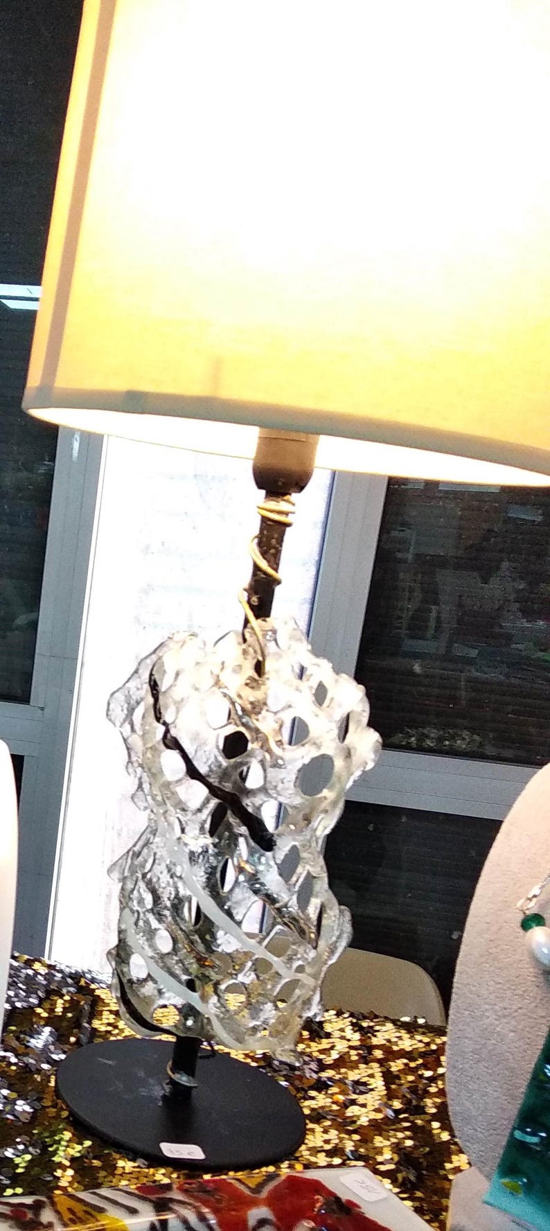 May include: A white glass lamp with a black base and a white fabric lampshade. The glass is shaped like a spiral with holes throughout. The lamp is sitting on a gold sequined table cloth.