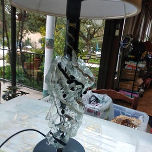 May include: A table lamp with a beige lampshade and a clear glass base with black accents. The base is made of many pieces of glass that are fused together to create a unique and modern design.