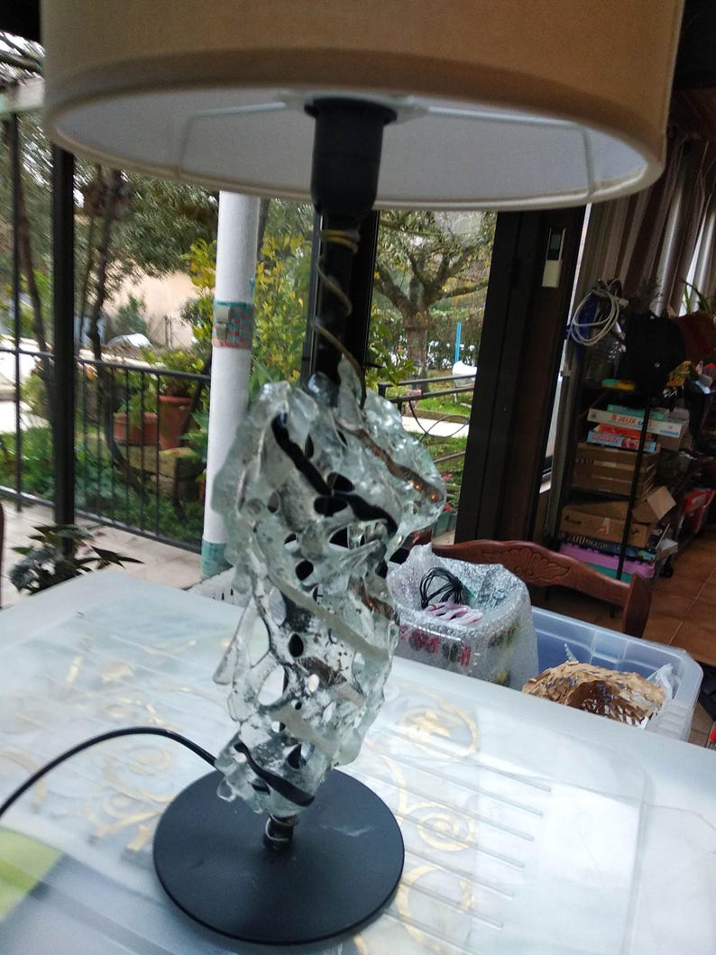 May include: A table lamp with a beige lampshade and a clear glass base with black accents. The base is made of many pieces of glass that are fused together to create a unique and modern design.
