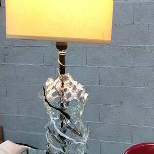 May include: A table lamp with a white fabric lampshade and a clear glass base with a black metal stem. The base is shaped like a twisted, abstract sculpture.