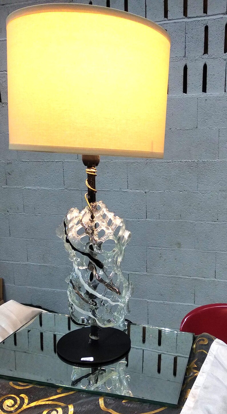 May include: A table lamp with a white fabric lampshade and a clear glass base with a black metal stem. The base is shaped like a twisted, abstract sculpture.