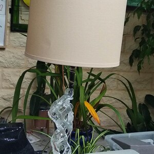 May include: A beige lampshade with a white fabric shade sits on a table. The lamp base is a clear glass sculpture with a black metal base.