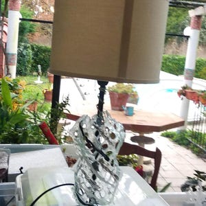 May include: A table lamp with a beige lampshade and a clear glass base with a decorative, intricate design. The base is made of glass and has a black metal stem.