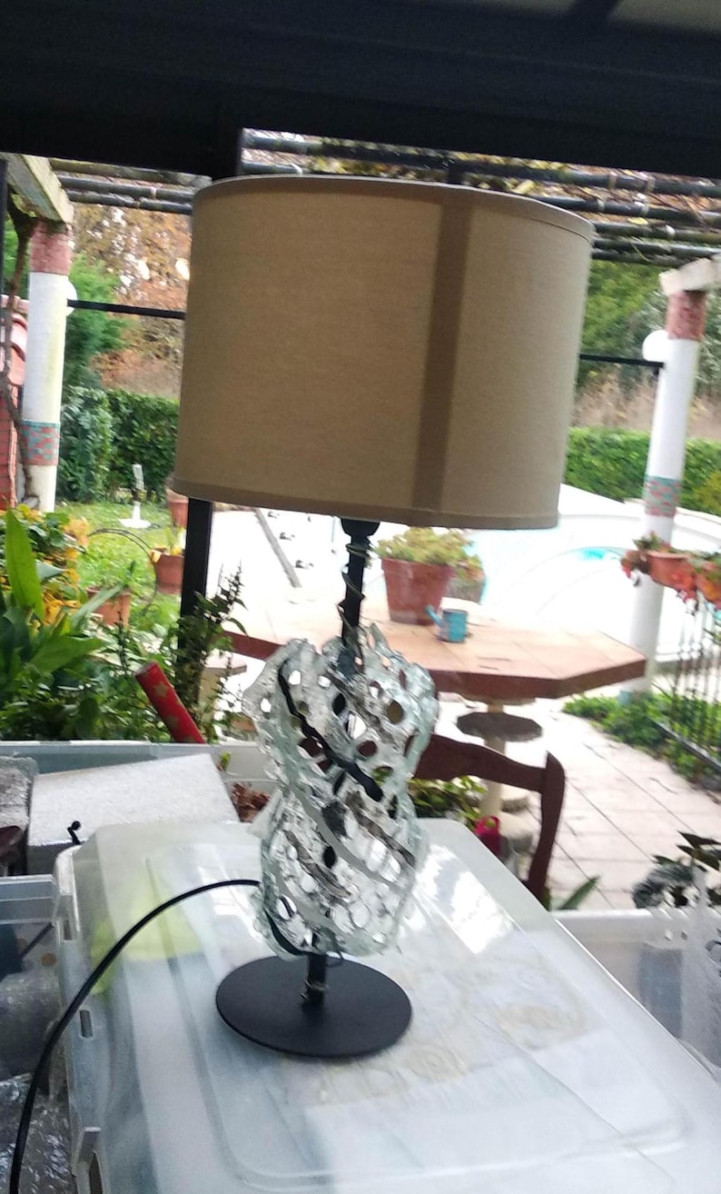 May include: A table lamp with a beige lampshade and a clear glass base with a decorative, intricate design. The base is made of glass and has a black metal stem.