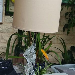 May include: A white lampshade with a glass base shaped like a tree branch. The lamp is on a white surface.