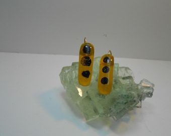 May include: A pair of handmade earrings. Each earring is a rectangular shape with a gold-colored base and black polka dots. The earrings are displayed on a light green, crystalline structure.