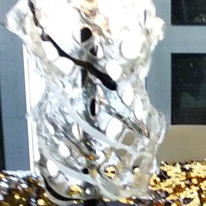 May include: A clear glass sculpture with a swirling, organic design. The sculpture is mounted on a black metal base and stands on a gold sequined surface.