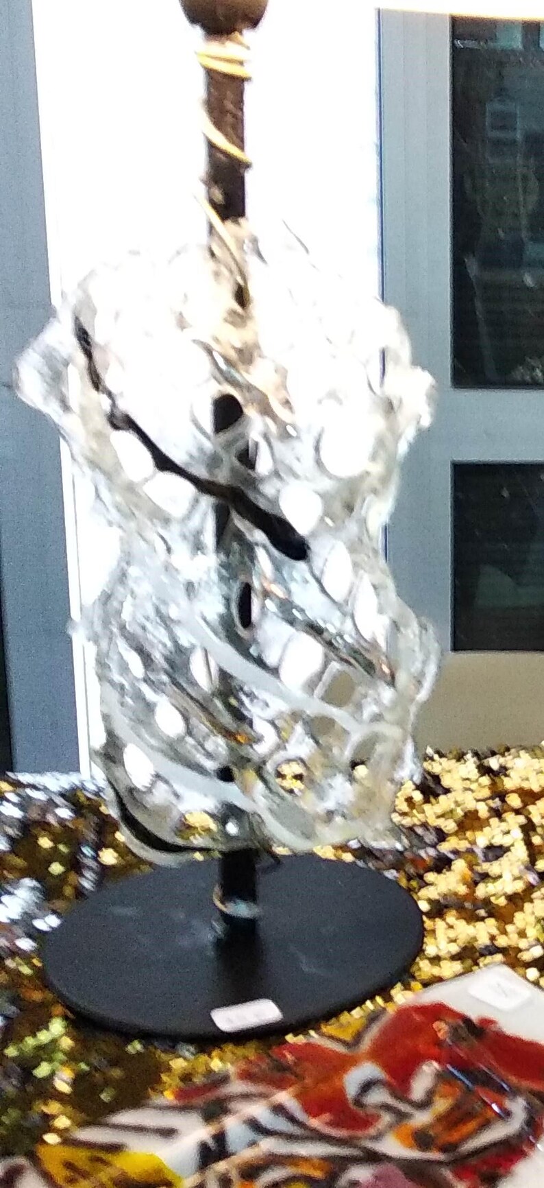 May include: A clear glass sculpture with a swirling, organic design. The sculpture is mounted on a black metal base and stands on a gold sequined surface.