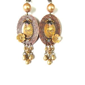 Peut inclure: Une paire de boucles d'oreilles pendantes avec des plaques ovales en métal cuivré aux motifs complexes. Chaque plaque présente un portrait central, une petite fleur dorée et un cristal clair. Trois perles dorées pendent en bas.