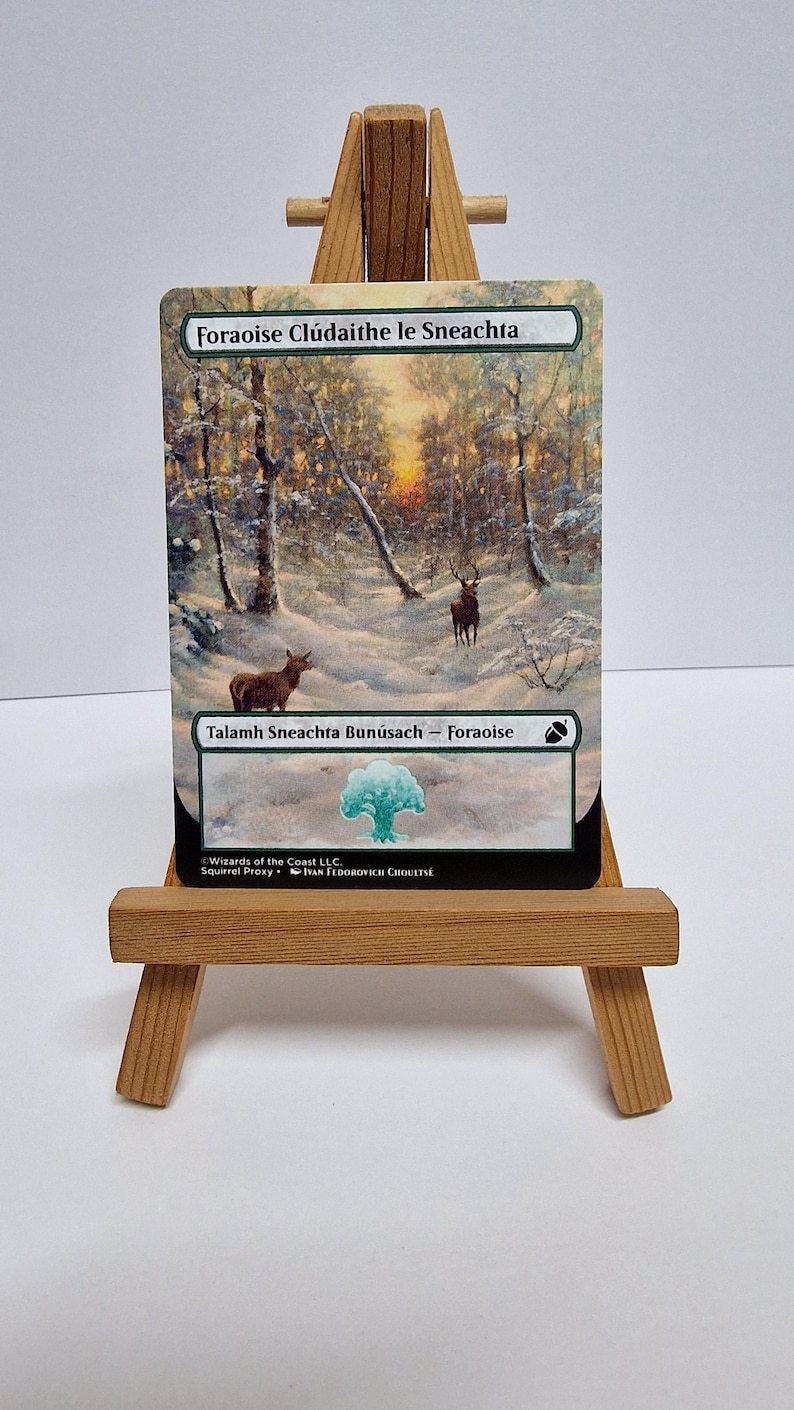 May include: A Magic: The Gathering card depicting a snowy forest with deer, displayed on a small wooden easel. The card's title is "Foraoise Cl&uacute;daithe le Sneachta" and includes text at the bottom. The card is displayed on a small wooden easel.