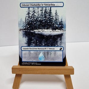 May include: A trading card depicting a winter scene with a lake and snow-covered trees. The card has Gaelic text. It is presented on a small wooden easel.