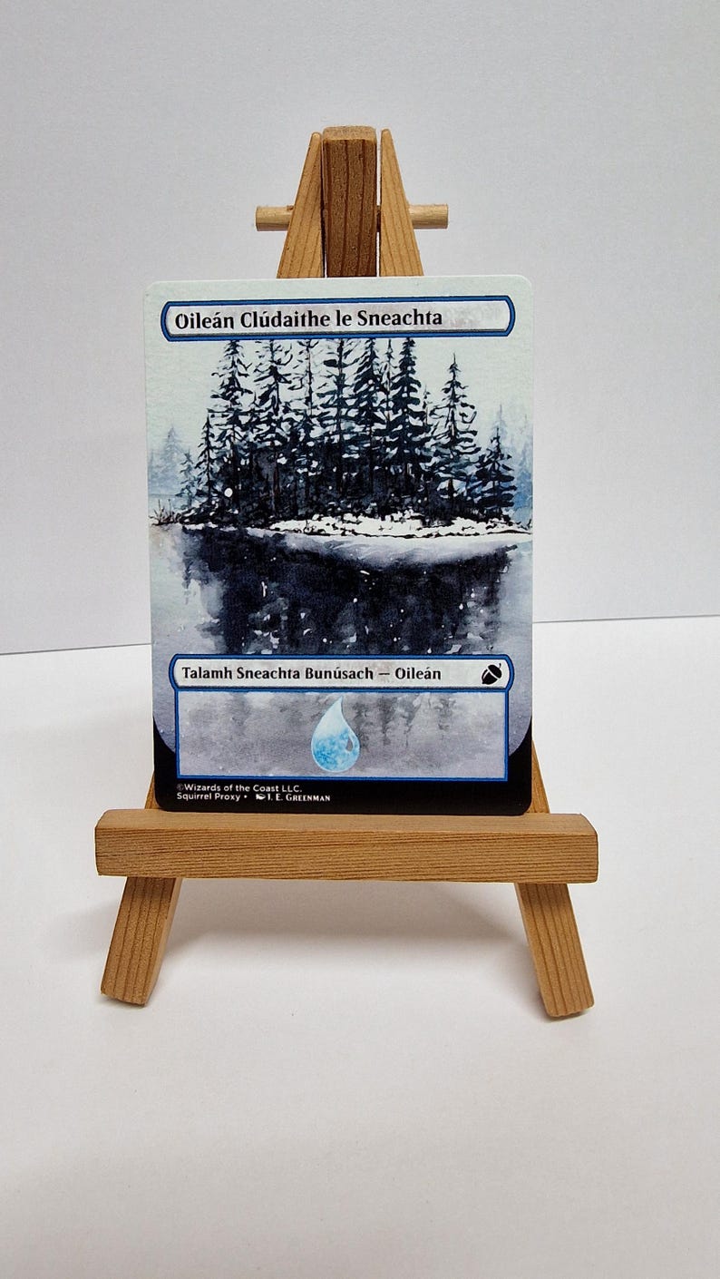 May include: A trading card depicting a winter scene with a lake and snow-covered trees. The card has Gaelic text. It is presented on a small wooden easel.