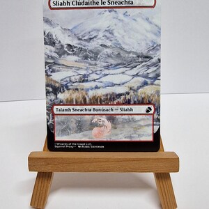 May include: A card with a snowy mountain scene, presented on a miniature wooden easel. The card includes Gaelic text, such as "Sliabh Cl&uacute;daithe le Sneachta" and "Talamh Sneachta Bun&uacute;sach - Sliabh". The image shows a mountain range covered in snow.