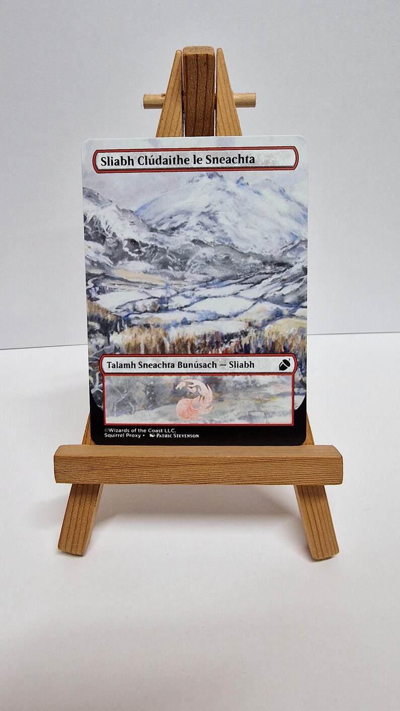May include: A card with a snowy mountain scene, presented on a miniature wooden easel. The card includes Gaelic text, such as "Sliabh Cl&uacute;daithe le Sneachta" and "Talamh Sneachta Bun&uacute;sach - Sliabh". The image shows a mountain range covered in snow.