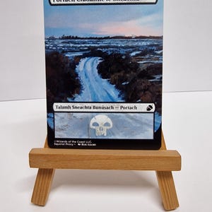 May include: A Magic: The Gathering card depicting a road through a snowy landscape under a cloudy sky. The card has Gaelic text and is presented on a small wooden easel.