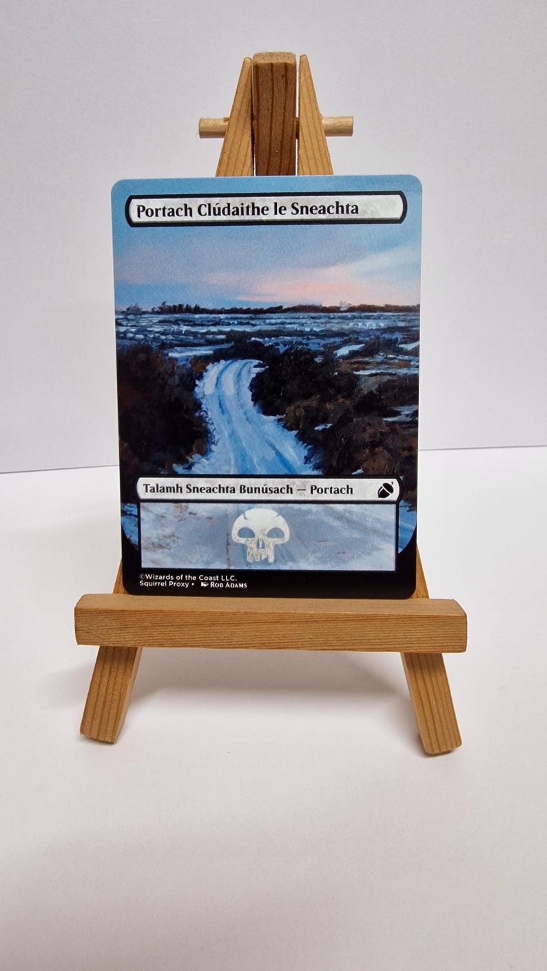 May include: A Magic: The Gathering card depicting a road through a snowy landscape under a cloudy sky. The card has Gaelic text and is presented on a small wooden easel.