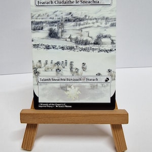 May include: A card depicting a snowy scene with a church and sheep. The text at the top says "F&eacute;arach Cl&uacute;daithe le Sneachta" and the bottom says "Talamh Sneachta Bun&uacute;sach - F&eacute;arach". The card is on a small wooden easel.