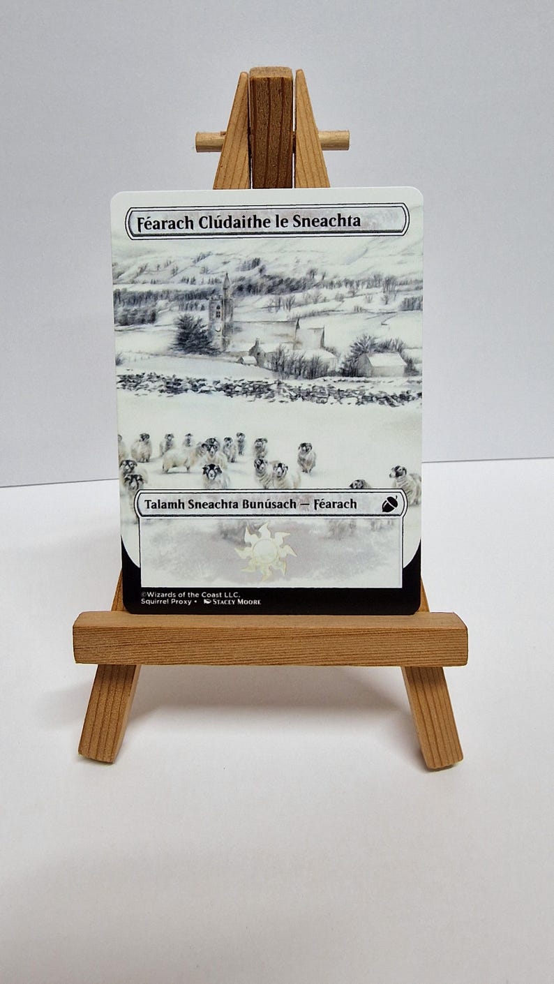 May include: A card depicting a snowy scene with a church and sheep. The text at the top says "F&eacute;arach Cl&uacute;daithe le Sneachta" and the bottom says "Talamh Sneachta Bun&uacute;sach - F&eacute;arach". The card is on a small wooden easel.