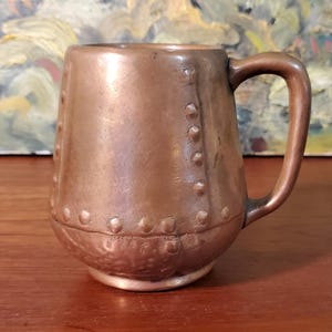 May include: A metallic copper-colored mug with a smooth, rounded body and a single handle. The exterior features a series of raised, circular rivet-like details arranged in vertical and horizontal lines, giving the appearance of joined metal plates.
