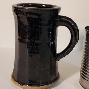 May include: A tall, dark brown, faceted ceramic mug with a glossy finish and a sturdy handle, positioned next to a standard, empty, silver-colored metal food can with horizontal ridges, providing a clear visual scale for the mug's height.
