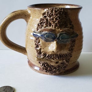May include: A tan speckled ceramic mug featuring a sculpted face with a textured beard, hair, and blue goggles. The mug has a curved handle and a smooth rim. A silver-colored US quarter coin is placed nearby for scale, showing the relative size.
