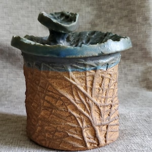 May include: A ceramic jar with a textured brown glaze and a blue glaze on the top. The jar has a unique lid with a raised design.