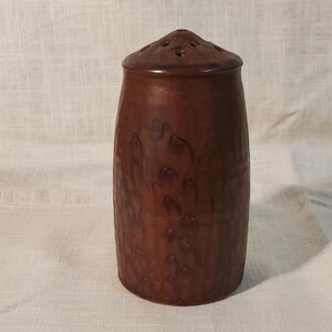 May include: A cylindrical brown ceramic shaker featuring a domed lid with multiple small holes at the top. The body of the shaker displays a mottled, drip-like glaze pattern in varying shades of dark and medium brown, creating a textured appearance.