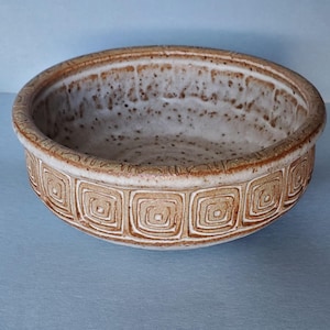 May include: A handcrafted ceramic bowl with a light brown exterior and a speckled interior. The bowl features a repeating square pattern around the rim. This decorative bowl is suitable for serving or display.