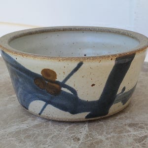 May include: A round ceramic bowl with a speckled off-white glaze. The exterior features a hand-painted dark blue abstract brushstroke design accented with small brown circular spots. The rim is unglazed, revealing the natural tan clay texture.