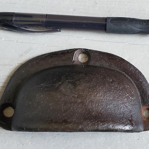 May include: A dark brown, semi-circular metal drawer pull with three holes for mounting. A black pen rests above the pull. The drawer pull has a rustic, aged appearance, suggesting it is vintage or antique. The pen is black with a rubber grip.