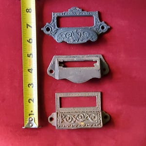 May include: Three antique metal drawer pulls with ornate designs. The top pull has a floral design, the middle pull is plain, and the bottom pull has a more intricate floral design.