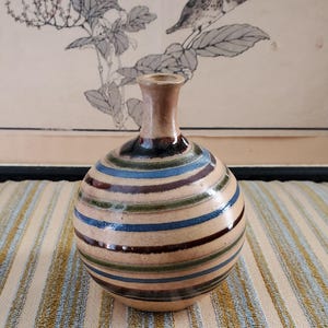 May include: A ceramic vase with a round body and a narrow neck. The vase is decorated with horizontal stripes in shades of brown, blue, and green. The background features a painting of a bird and foliage.