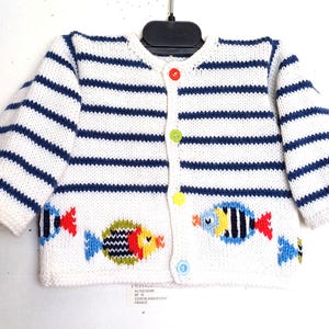 Könnte beinhalten: Weiß-blau gestreifte Baby-Strickjacke mit bunten Fischstickereien. Der Cardigan mit Knöpfen hat einen runden Ausschnitt und lange Ärmel. Die Knöpfe sind rot, grün, gelb und blau.