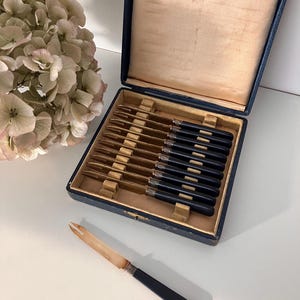 May include: A set of antique fruit knives in a blue box. The knives have black handles and gold-toned blades. The box is lined with beige fabric and has a handwritten inscription. One knife is outside the box.