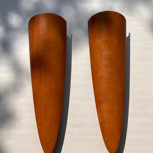 May include: Two orange, cone-shaped wall sconces. The sconces have a smooth, textured surface and a dark shadow on the back. The sconces are mounted on a white wall. The sconces are a decorative lighting fixture.