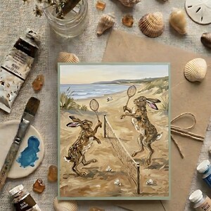 Peut inclure: Une impression d'art rectangulaire représentant une peinture de deux lièvres bruns jouant au badminton sur une plage de sable avec un filet, des raquettes et des volants. La scène inclut un océan bleu et des dunes en arrière-plan.
