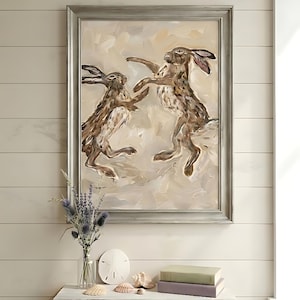 May include: A framed oil painting featuring two brown hares in a dynamic jumping pose against a neutral, textured beige background. The artwork is enclosed in a weathered grey wooden frame, showcasing expressive brushstrokes and earthy tones.