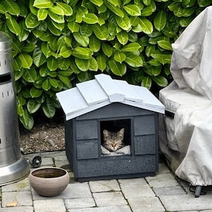 May include: A gray cat house with a white roof and a tabby cat sleeping inside. The cat house is on a patio with a silver propane heater and a gray covered chair.