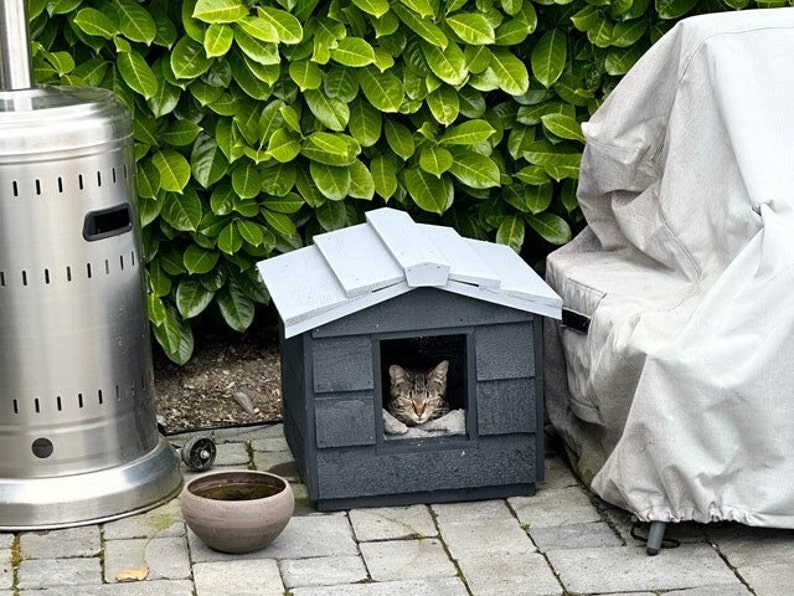 May include: A gray cat house with a white roof and a tabby cat sleeping inside. The cat house is on a patio with a silver propane heater and a gray covered chair.